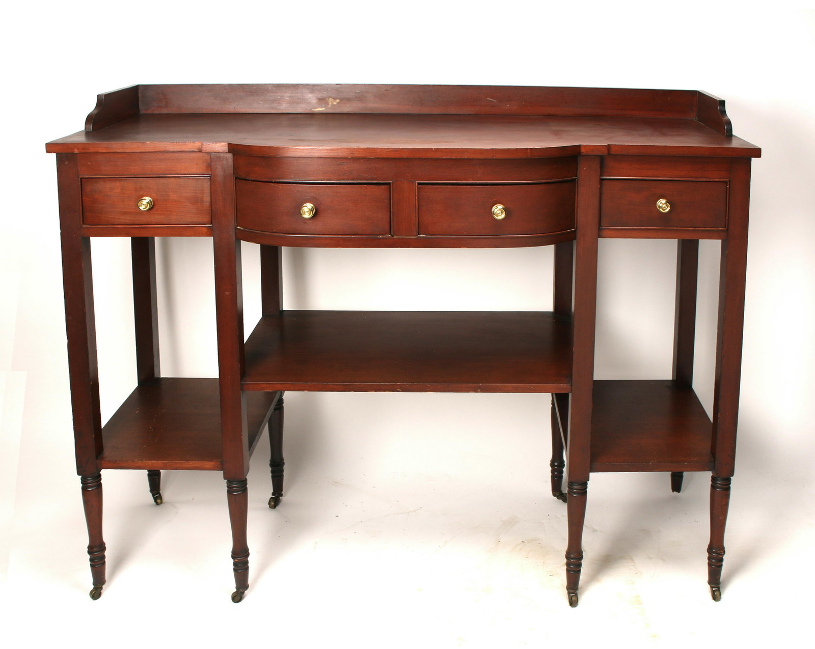 FEDERAL STYLE MAHOGANY SIDEBOARD: Sideboard with shaped front, eight turned legs with casters, four drawers with brass pulls, and two tier bottom shelf area. Central bow front. Has the advantage of having shallow depth with a lot of s