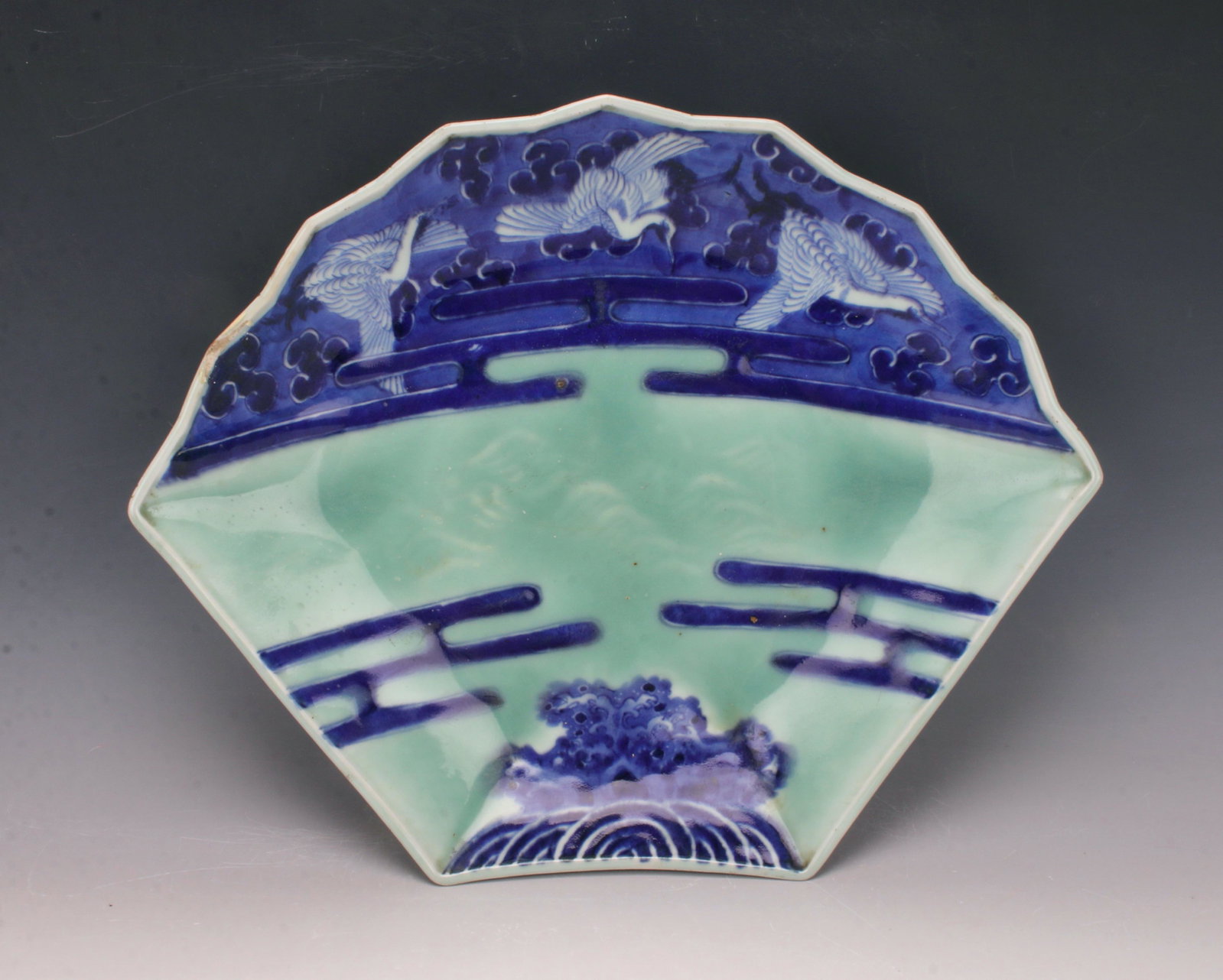 KOREAN FAN SHAPE CHARGER: Cranes fly among clouds. A subtle mountain scene shows through the celadon central area. Outer rim has mountains. Marked on the bottom. Provenance: