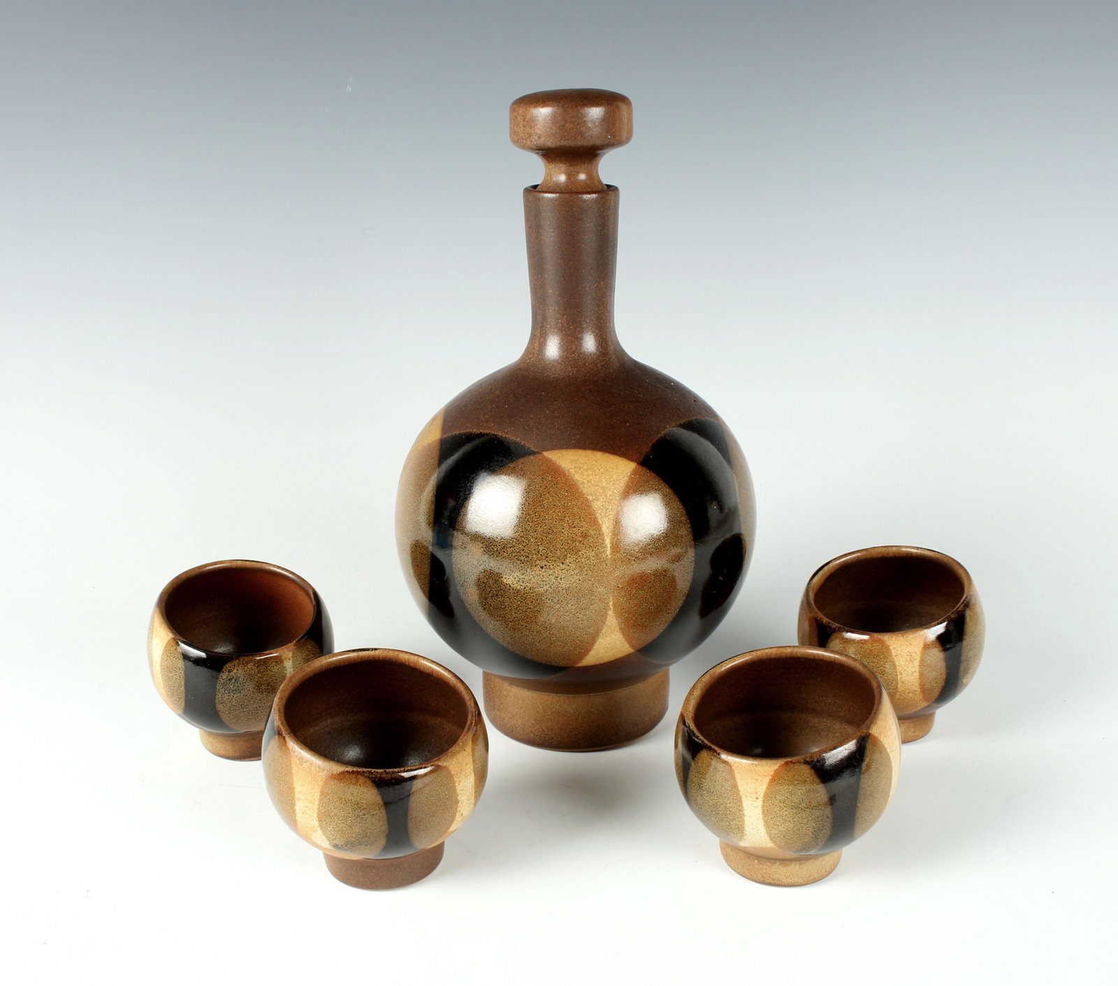 1960S CERAMIC DECANTER 4 CUPS SAKE SET: Combining op art and earthtones in a stylized Mid Century sake set. Glazed in overlapping circles for distinctive 1960’s vibe. Four rounded cups are set on an angle. Unmarked. 5" X 10"