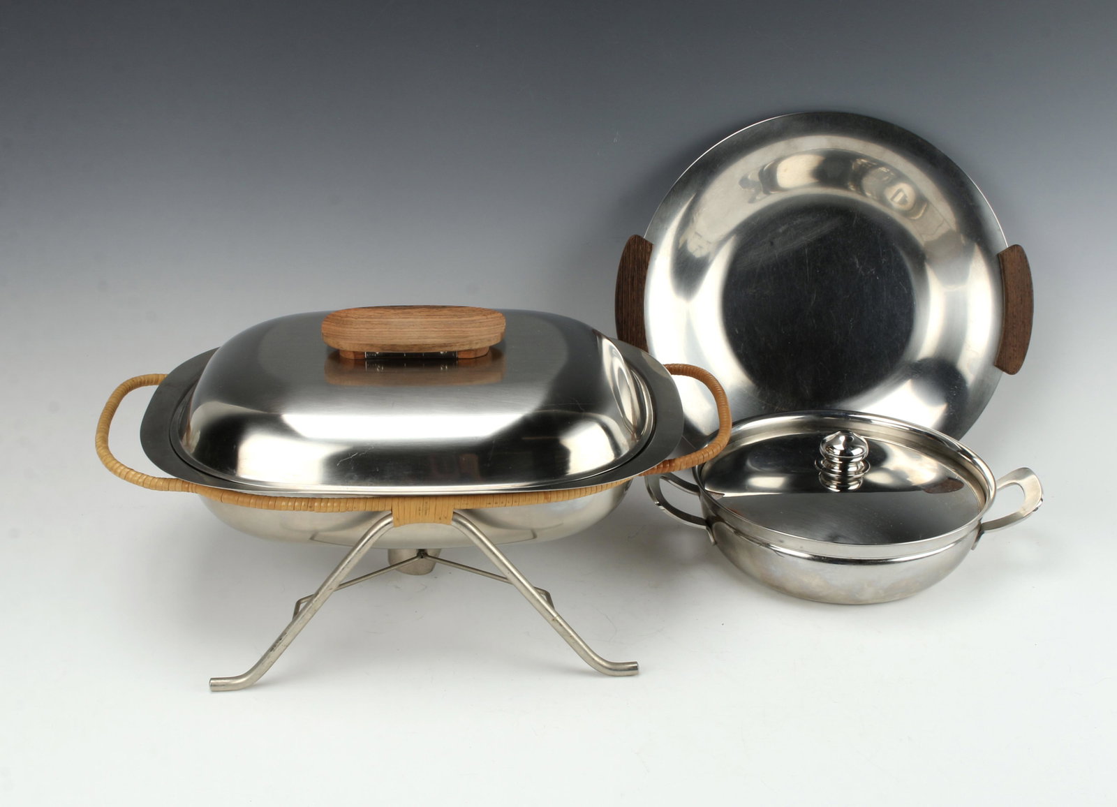 3 MID CENTURY SERVING PIECES: Lidding casserole dish is marked Jero Zilmeta. Stainless chaffing dish with a teak handle and wicker wrapped frame with warmer. Round tray with wood handles. 14 1/2" X 7 1/2" $100 - $200<