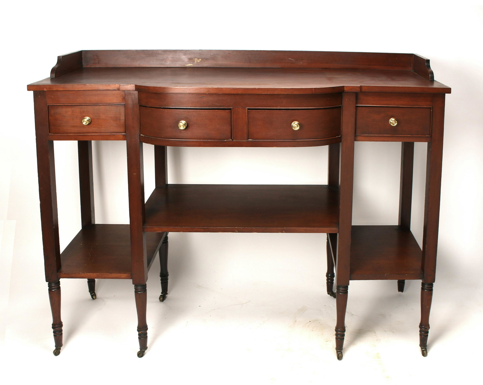 FEDERAL STYLE MAHOGANY SIDEBOARD: Sideboard with shaped front, eight turned legs with casters, four drawers with brass pulls, and two tier bottom shelf area. Central bow front. Has the advantage of having shallow depth with a lot of s