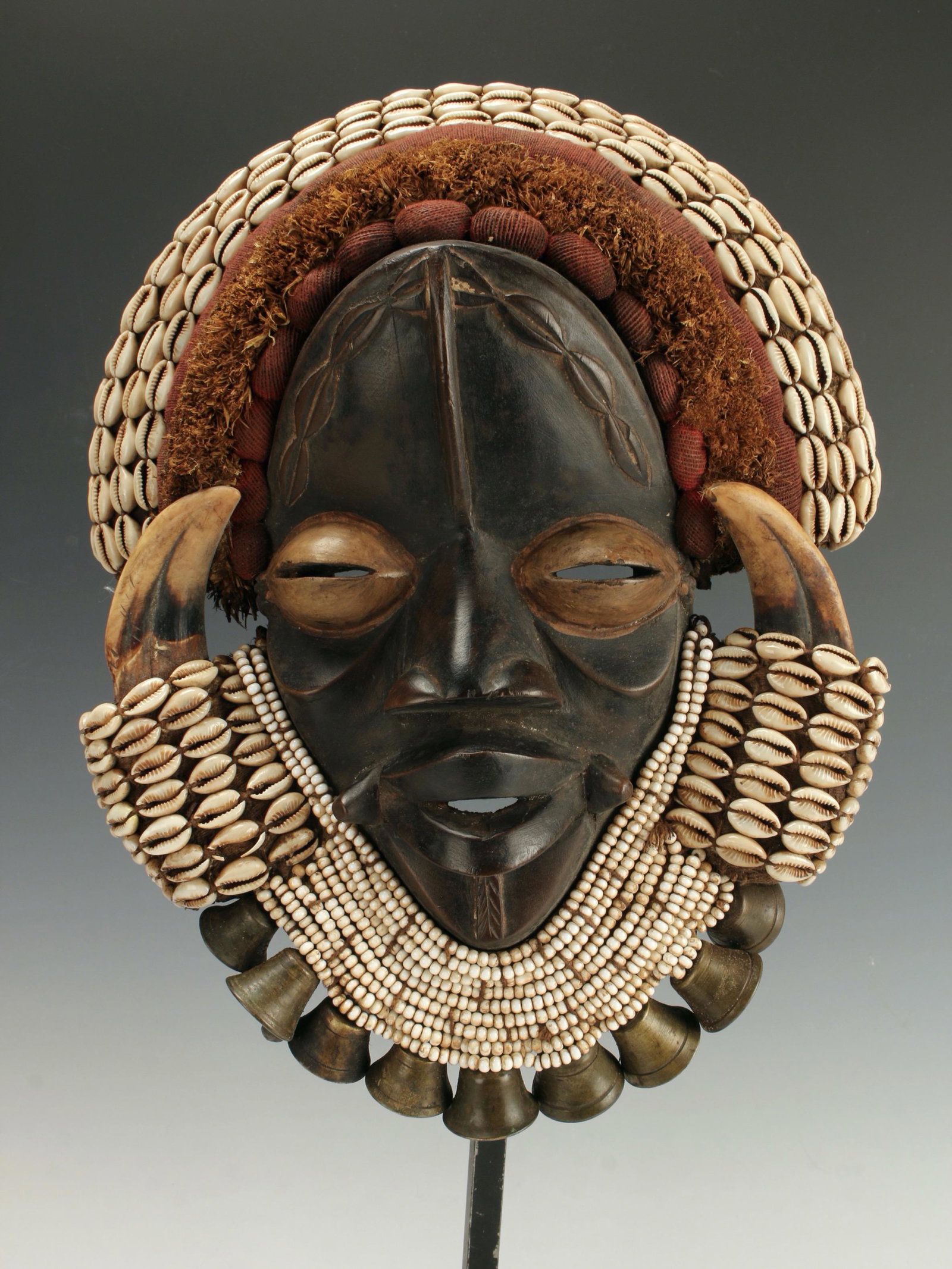 DAN GUERE MASK W COWRIE & BELLS IVORY COAST: Impressive mask. Made from wood, wild boar horn, cowrie shells, beads, textiles, raffia, and bronze bells. On custom metal and wood stand. 23" X 10" $300 - $500 Please contact us for