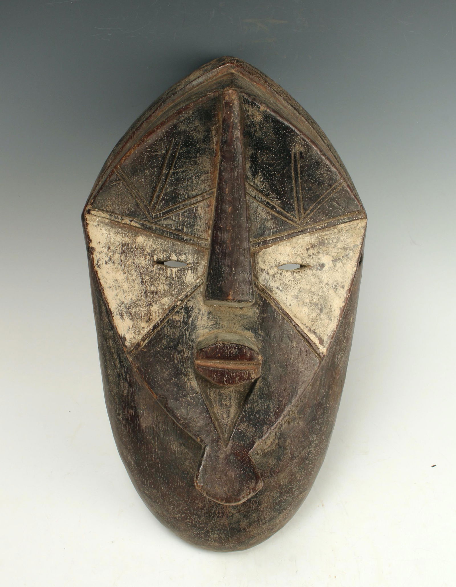 LUALUA LWALWA MASK CONGO: Black mask with white mask at the eyes. Incised geometric patterns on forehead. Protruding triangular nose and lips, shaped chin. Concave face. Wide frame around face and down neck. 15" X 8"