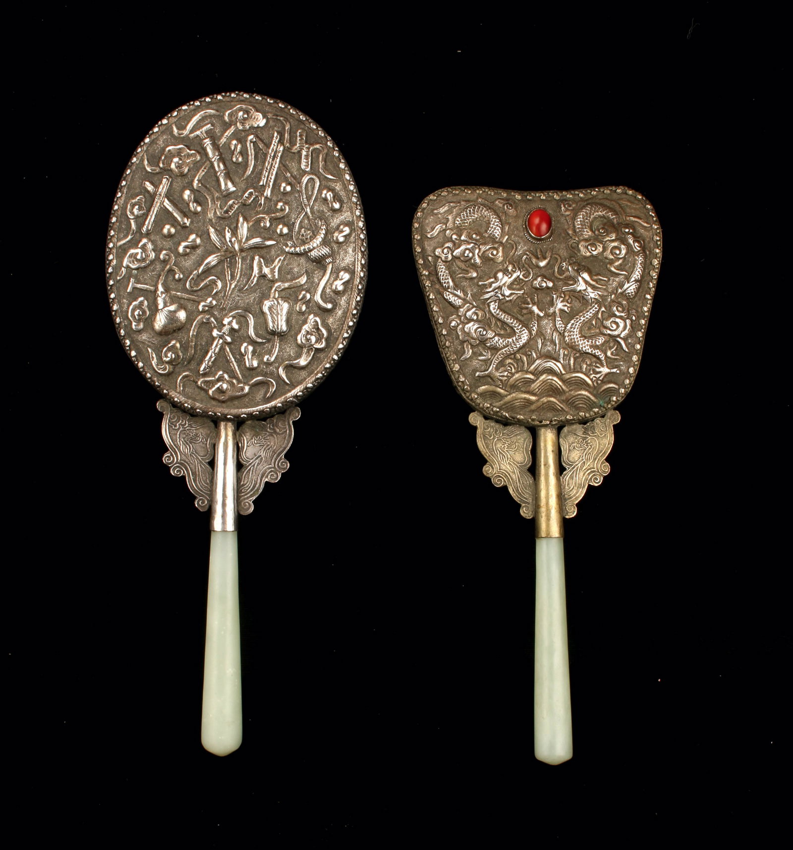2 CHINESE REPOUSSE HAND MIRRORS: With repousse designs. One is fan shaped, has facing dragons and a bezel set carnelian stone. The other is oval with repousse designs of scholars items. The stone handles are joined to the mirror with