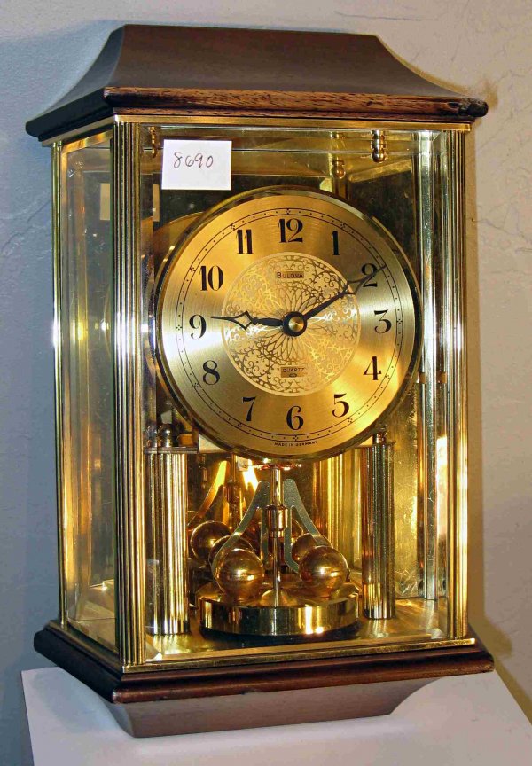 Bulova wall clock: A 20th century Bulova quartz clock presented within a bevel glass, gold toned metal and walnut bound wall case with torsion type pendulum and gold toned dial.
