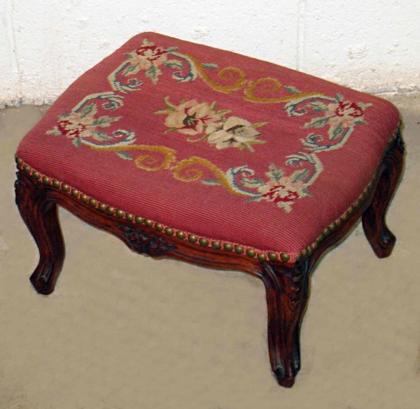 Needlepoint Covered Footstool: A carved wood framed footstool with needlepoint cover with a red background.