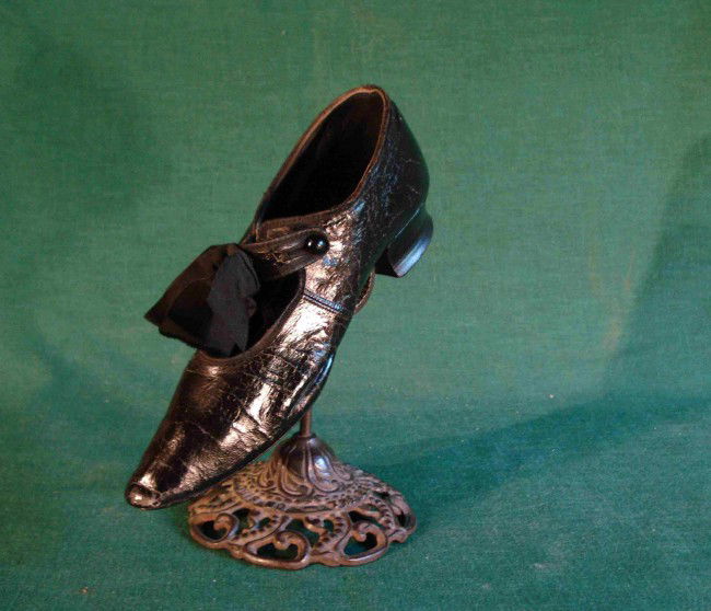 Shoe saleman's display stand: Here's how they sold shoes 100 years ago. An authentic 19th or early 20th century black leather shoe with a single button clasp and satin bow on a cast iron by the Belle Hickey Mfg Co of St. Louis.