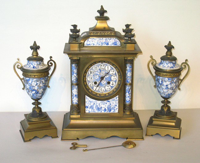 French Garniture Clock Set: A French made clock set featuring a brass mantel clock set with porcelain panels and matching brass urns with matching porcelain.