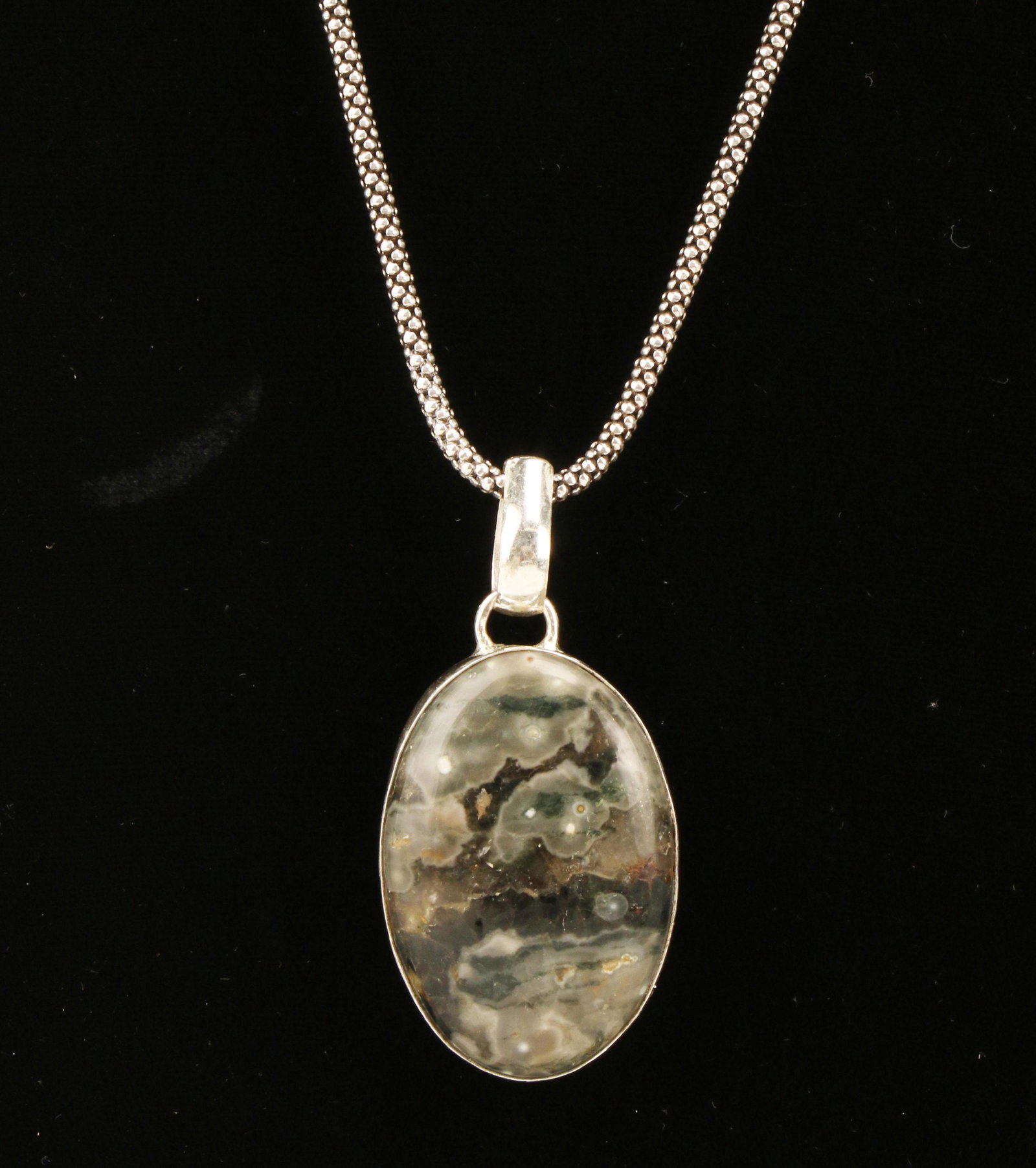 BLACK NATURAL STONE & STERLING PENDANT ON CHAIN: BLACK NATURAL STONE & STERLING PENDANT ON CHAIN Oval cabochon stone with clouds of grays and blacks. Possibly snow flake jasper. 18 1/2” chain, weight