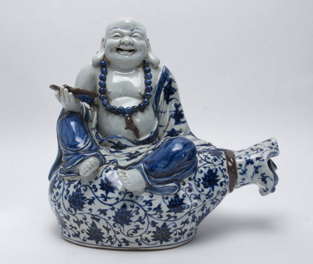 LAUGHING BUDDHA IN BLUE AND WHITE: Laughing Buddha In Blue And White: A laughing blue and white Buddha of safe travels sits on a hemp sack for protection and holds a bat in his left hand. The sack in painted with leaves and swirling v