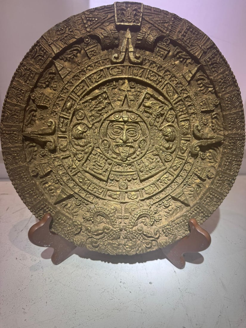 Bronze Plaque After the Aztec Calendar (Sun Stone): The bronze surface displays a warm, aged patina, consistent with cast decorative bronzes. Mounted on a wooden display stand. Likely a 20th-century decorative or educational work inspired by