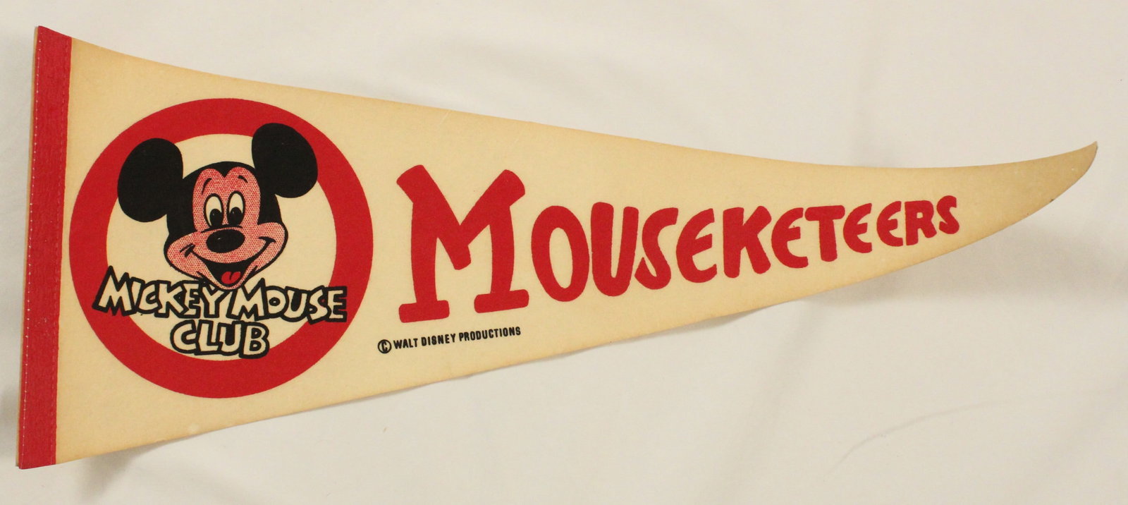 A Mickey Mouse Club Pennant: This is a red and cream colored pennant featuring the Mickey Mouse Club logo and "Mousketeers " in large red font. It is in good condition with some bending to the pennant and pin marks along the edge