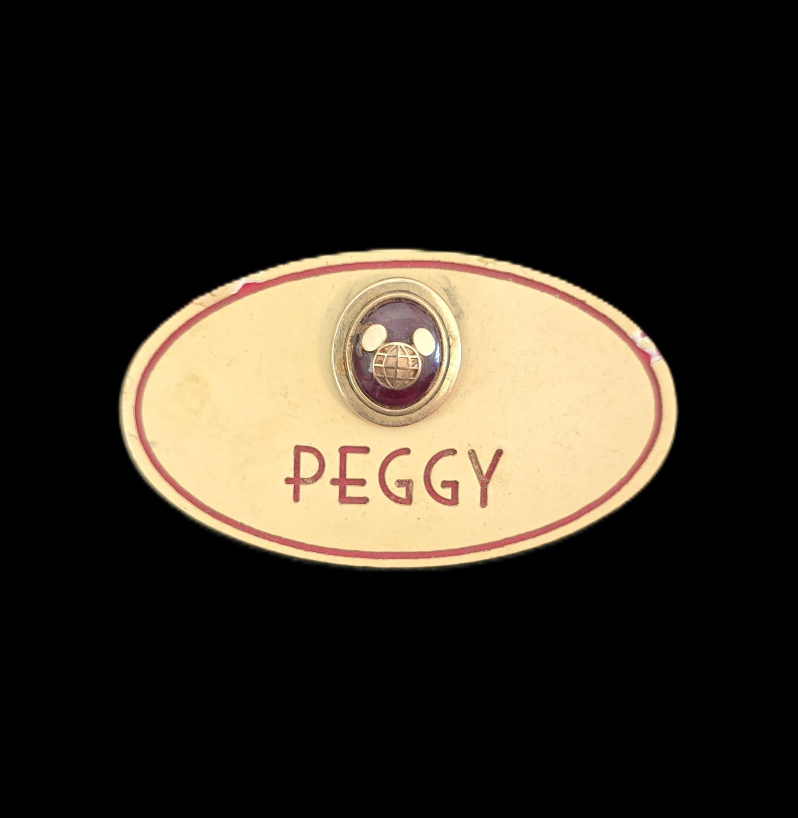 A Walt Disney World Cast Member "Peggy" Name Tag with Ruby 5-Year Pin.: (Disney, 1990s) This is a cast member name tag for Peggy. It includes the ruby 5-year award pin. The pin features a mickey head globe icon over a large ruby oval in a gold colored frame. The name badg