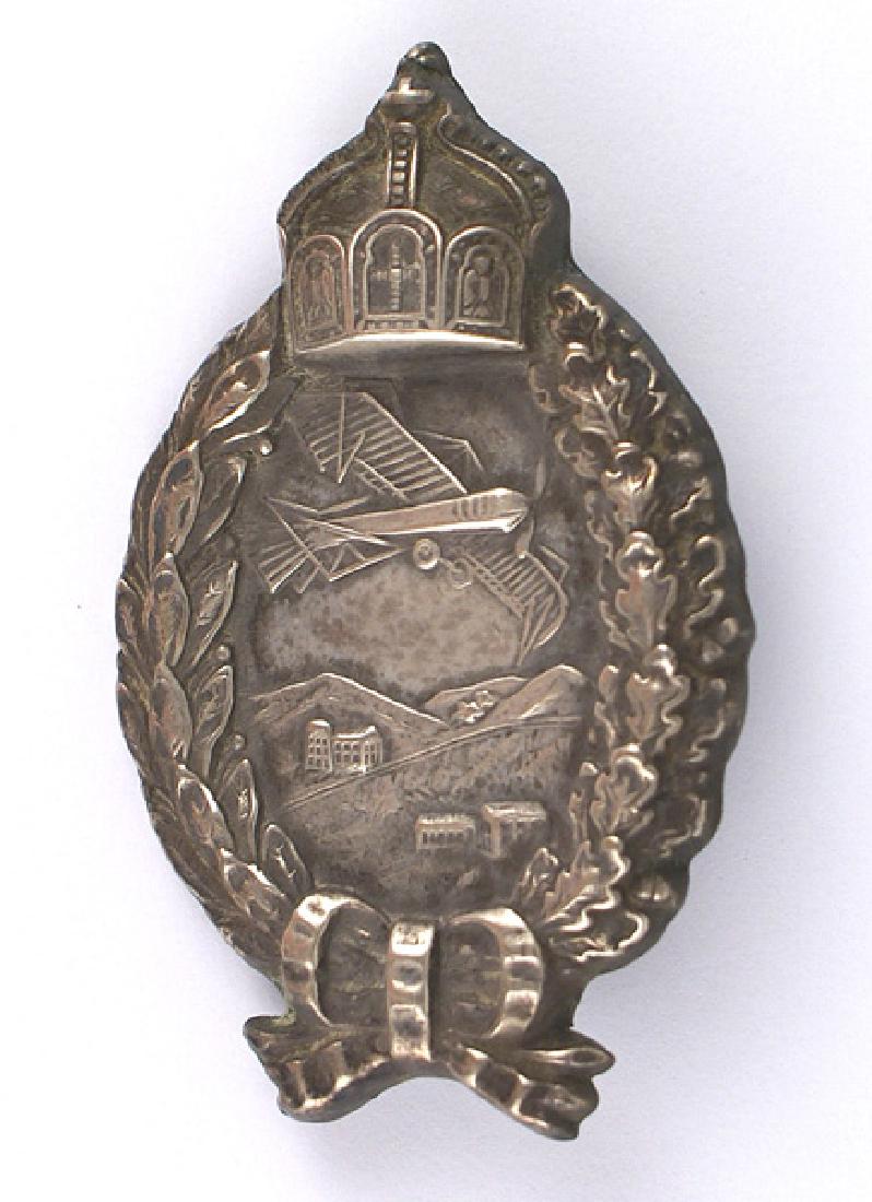 Prussian WWI Pilot Badge Medal: B-044 Silver Prussian WWI Pilot's Badge. View of an early-type aircraft flying over a mountain village is enclosed within a high relief oak/laurel leaf wreath with a large Hohenzollern Crown at the to