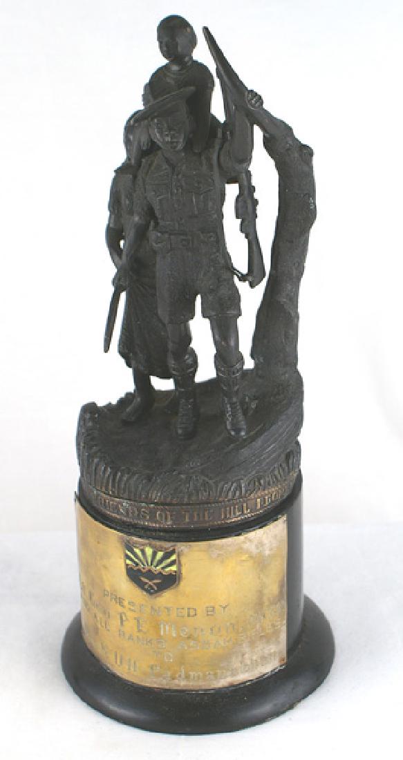 Statue of Gurkha presented ASSAM RIFLES: G-018 Darkened 8" metal statue of a Gurkha, with rifle and kukri, carrying his son, next to his wife. 3 1/2" round wood base has a full brass plate with an enameled unit shield and inscribed "Presente
