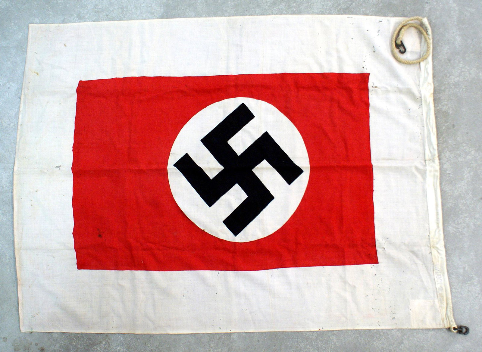 German WWII Harbor Pilot flag: H-066 Harbor Pilot's flag. Printed swastika roundel on a 25" x 37" red field with a white border. Rope sleeve is marked "Lotsenflg. 100/140". Metal hooks attached to ropes on each end. Overall, 40" x 