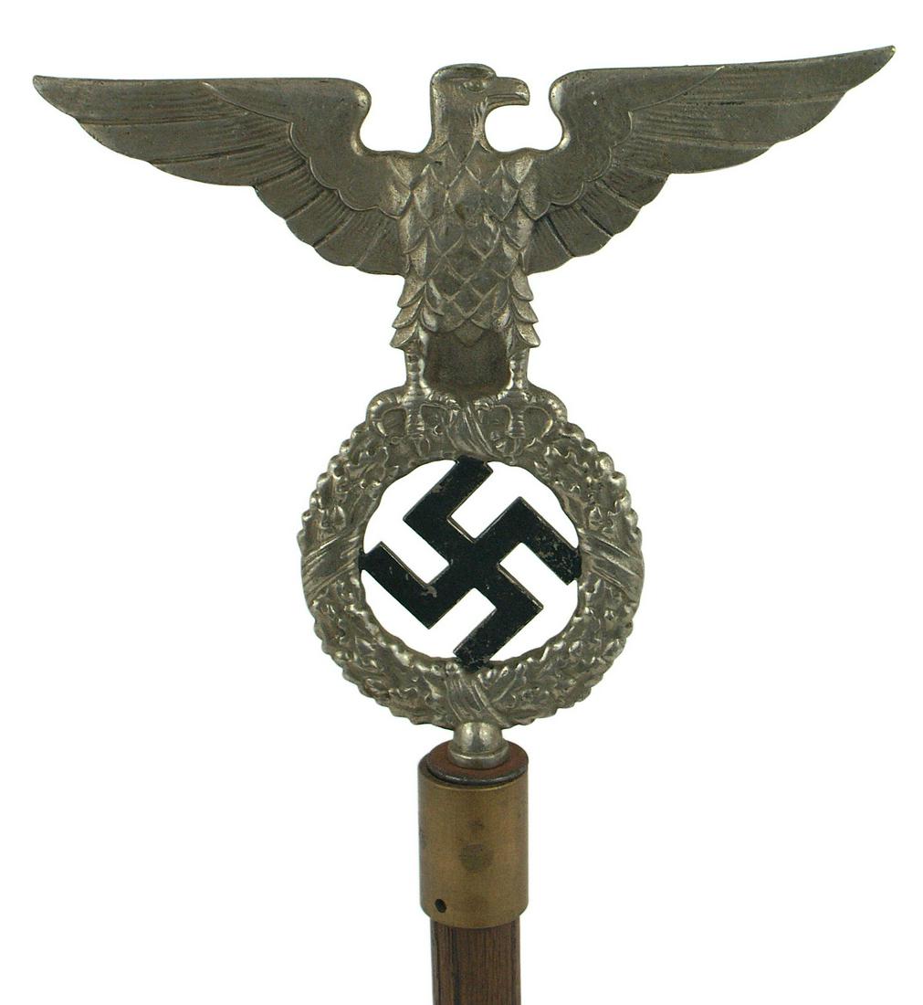 German WWII NSDAP nickeled pole top: H-067 N.S.D.A.P. nickeled pole top. 3 1/2" x 8 1/4" eagle perched on a large open oak leaf wreath holding a black swastika, overall 7" tall. Mounted on a 9" wood post with a metal socket and round woo