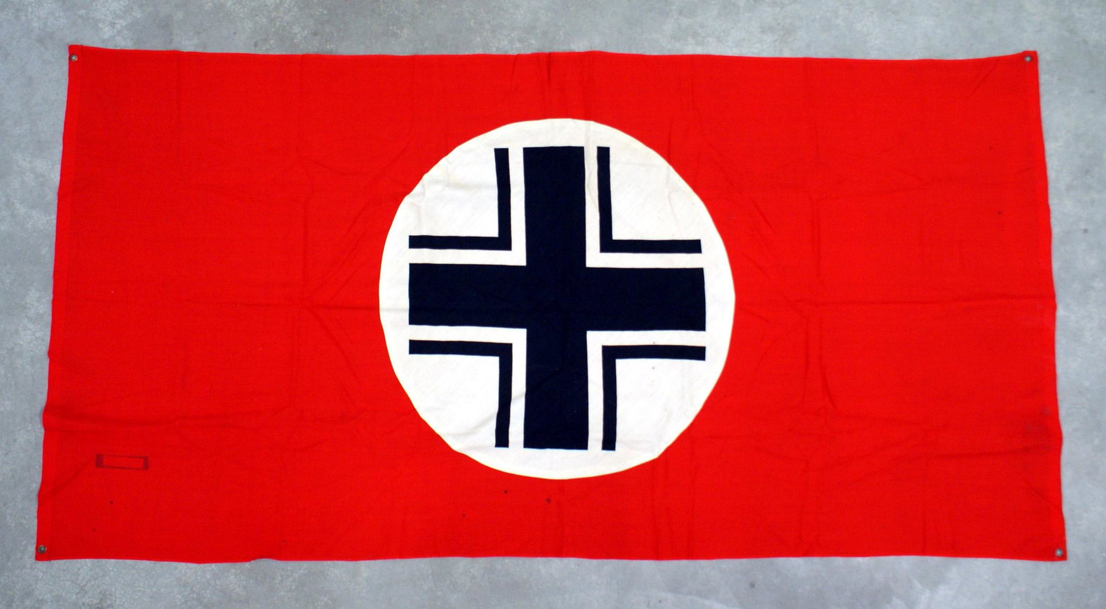German WWII military ID vehicle flag: H-077 German WWII military identification (from the air) vehicle flag. 40" x 77" red field with a 26 1/2" diameter white roundel holding a black Balkan Cross. Brass grommet in each of the corners. II+