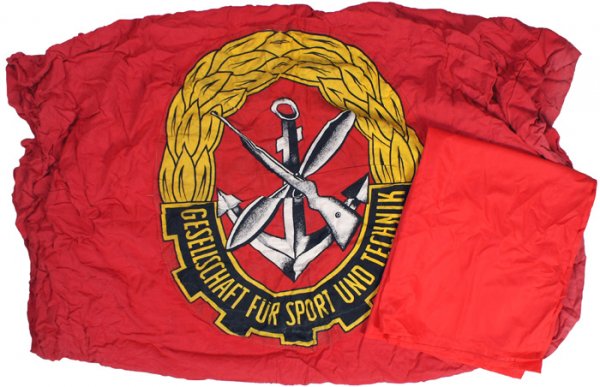 Lot of 2 East German Flags: H-142 - Lot of 2 East German Flags: (1) Red with a large sewn 25" center with an anchor, crossed rifle, propeller and "GESELLSCHAFT FUR SPORT UND TECHNIK" and gold wreath. With a sewn pole sleeve. 44"