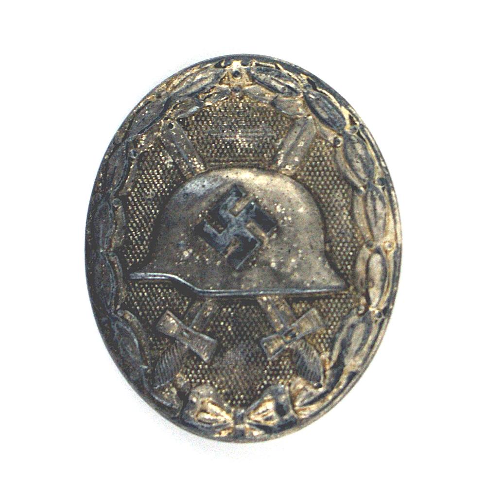 German WWII Silver Wound Badge: B-054 German WWII Silver Wound Badge. Relief swastika on helmet with crossed swords within a silvered oak/laurel leaf wreath. Age toned. II