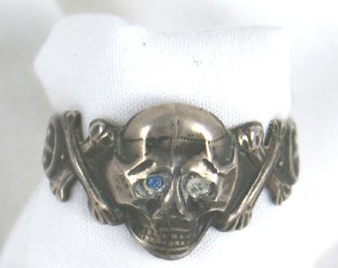 Private purchase German SS ring: H-118 Private purchase SS ring. Large skull with pronounced cheeks and jaw. One of two blue eye stones remains. Crossbones and floral work on sides. Inside band is marked "900". Shows age and wear. Ab