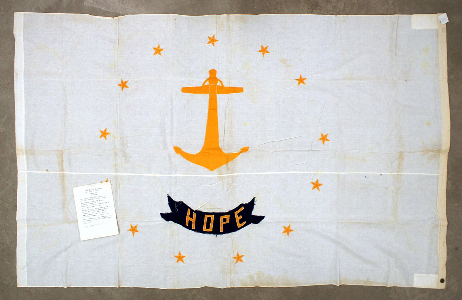 US State flag of Rhode Island: K-063 State flag of Rhode Island. White field with a large gold anchor surrounded by 13 separately sewn gold stars. Blue panel with "HOPE" in gold letters. Aged foxing on the white field. One grommet 