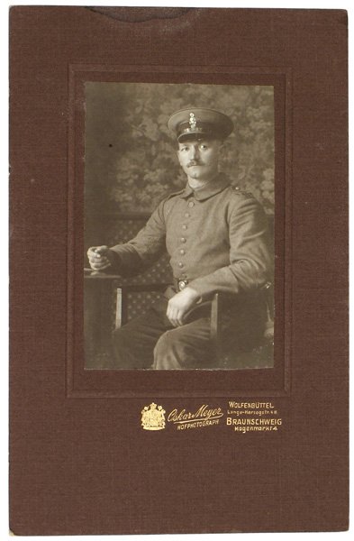 German WWI photo 17th Brunswick Hussars: L-1 Portrait of a seated member of the 17th Brunswick Hussars. Figure is wearing the wartime uniform of “feldgrau” and peaked cap with the skull device. In a mat frame. Gilt embossed logo of photo