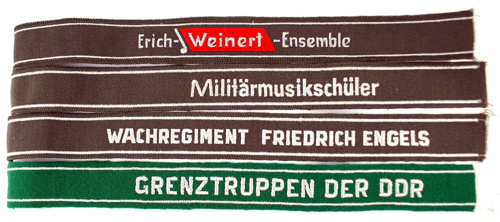 4 East German cufftitles DER DDR: H-139 Lot of 4 East German cufftitles. Includes: (1) "WACHREGIMENT FRIEDRICH ENGELS". (2) "MILITARMUSIKSCHULER". (3) "Erich - Weinert - Ensemble". And (4) "GRENSTRUPPEN DER DDR". Excellent I