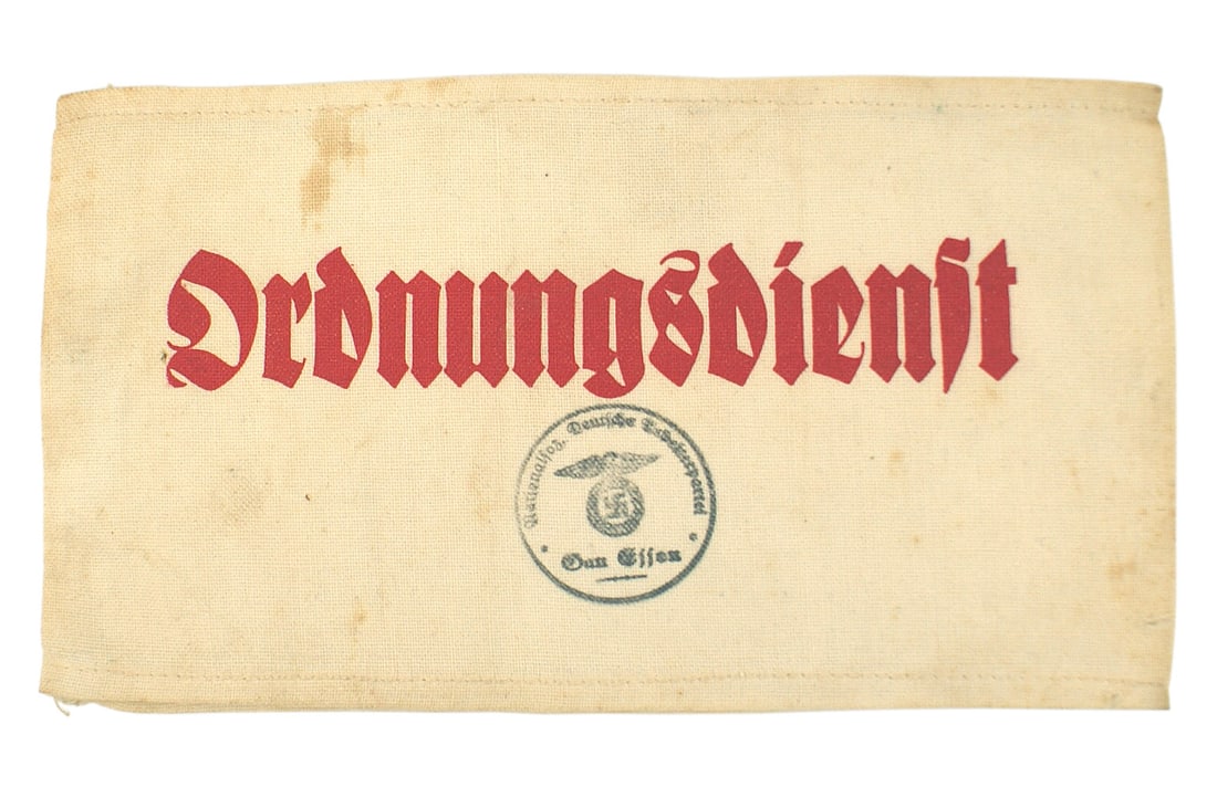 German WWII Military Police Service armband: H-016 Third-Reich red printed "Ordnungsdienst" (Military Police Service") armband. Also with a Party inkstamp for "Gau Essen". All on a white linen band. Some age foxing. Scarce. II