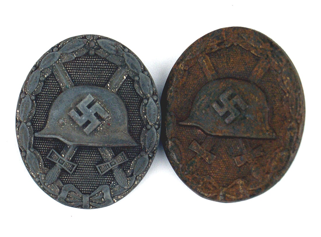 2 German WWII Wound Badges: B-050 Lot of 2 German WWII Wound Badges: (1) Relief swastika on helmet with crossed swords on a pebbled background. Oak leaf wreath border. Silver specks indicate the finish when issued. A solid, gray