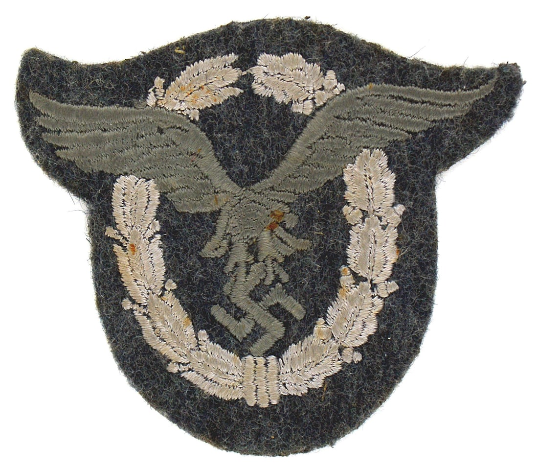German WWII Luftwaffe cloth Pilot Badge: B-067 German WWII Luftwaffe cloth Pilot's Badge. Embroidered silver-gray flying eagle/swastika within a light gray laurel//oak leaves wreath on a blue-gray background. A stiff backing. I