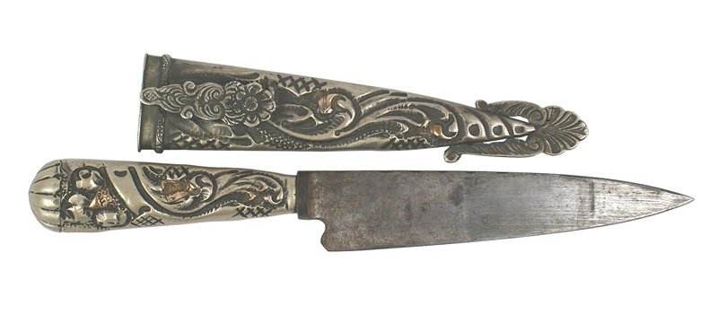 Argentine gaucho knife Circa 1930: D-44 Argentine "gaucho" knife. Relief silver/gilt floral work on the age toned decorative grip and scabbard. The 4 3/4" gray blade shows some wear, but "MINUS RIA..." is visible. Circa 1930. About II