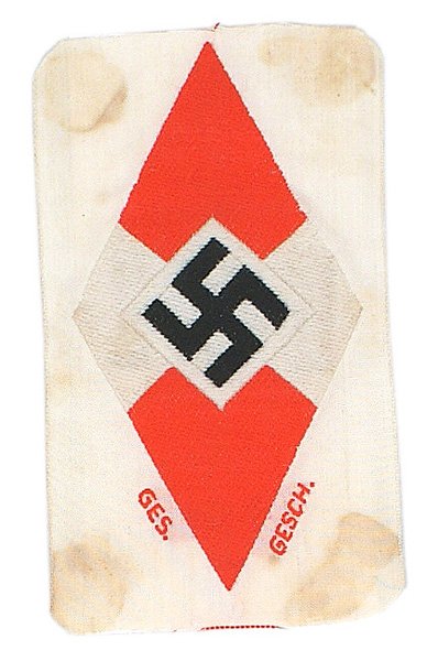 German WWII Hitler Youth diamond: H-166 Hitler Youth member's bevo embroidered sleeve diamond. Tilted black swastika on red/white diamond. Paper "RZM" tag. Uncut - marked "GES. GESCH". White cloth background is stained. II