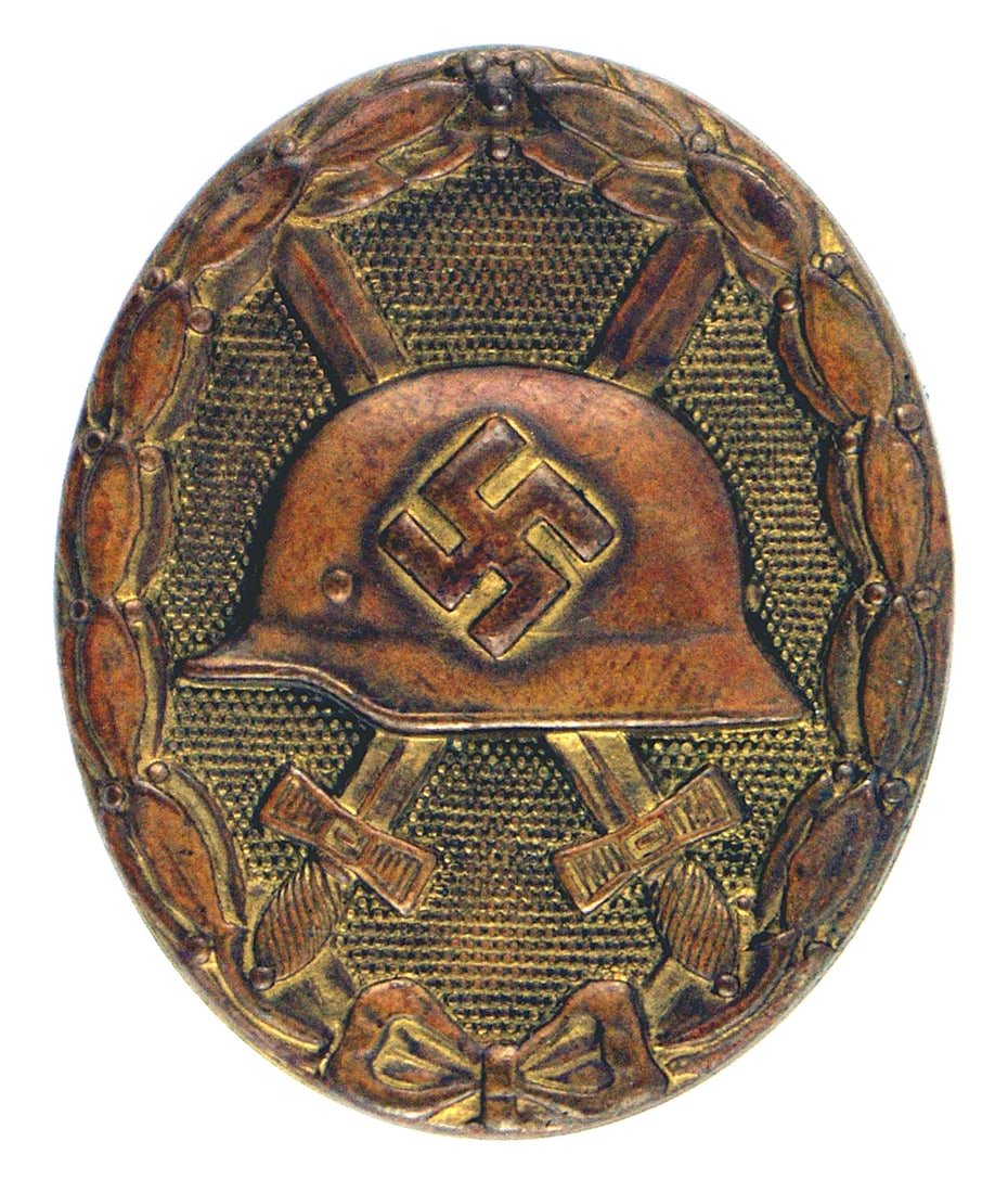 German WWII Wound Badge Gold: B-066 German Third Reich Wound Badge - Gold. Heavy brass type. Shows some gilt residue. Relief helmet with swastika and crossed swords on a stippled background within an oak leaf border wreath. II