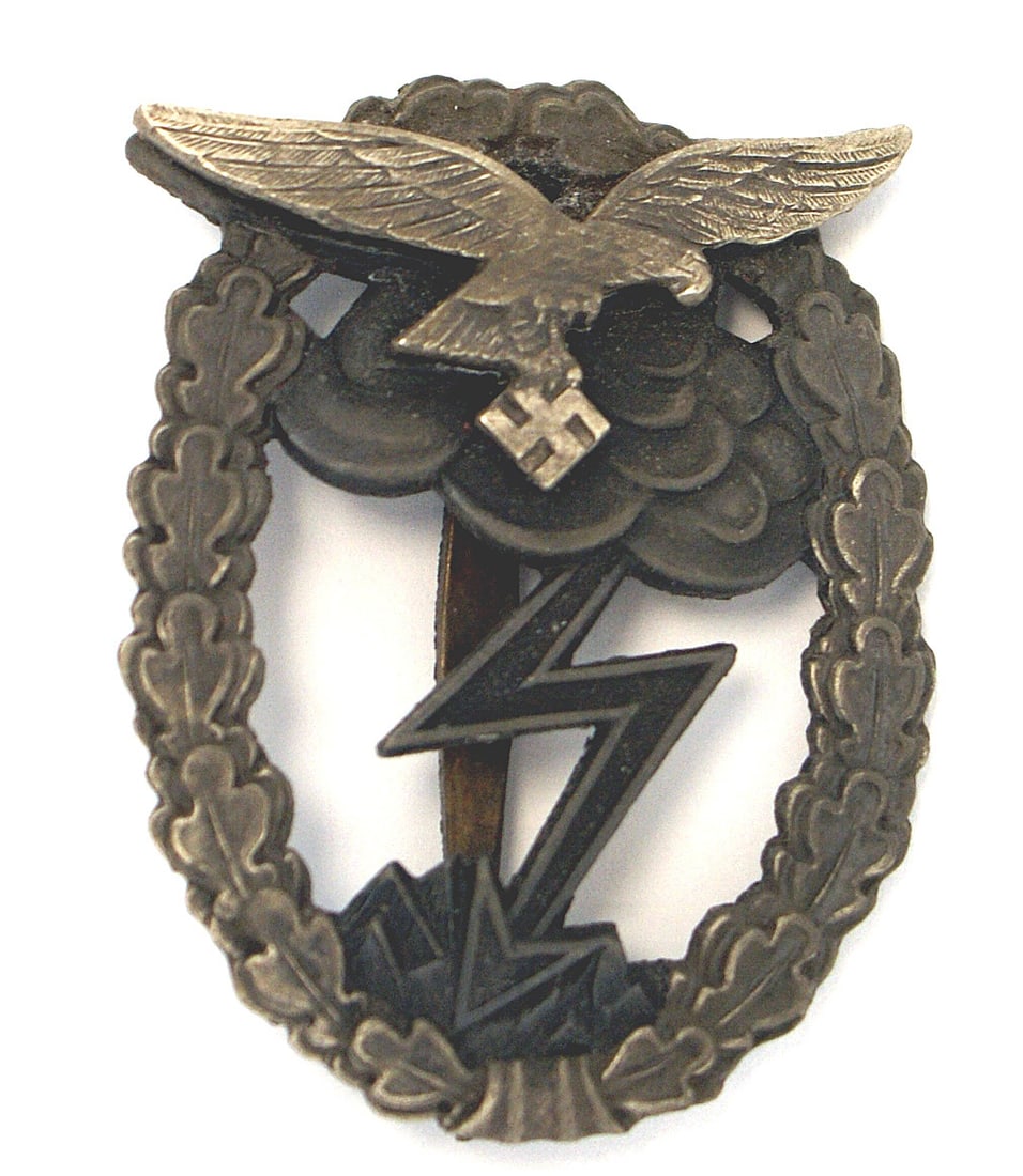 WWII Luftwaffe Ground Combat Badge: B-096 German WWII Luftwaffe Ground Combat Badge. Separately applied darkened silvered flying eagle/swastika on a gray metal wreath with clouds/lightning bolt. Slightly dished reverse marked "M.u.K.". 