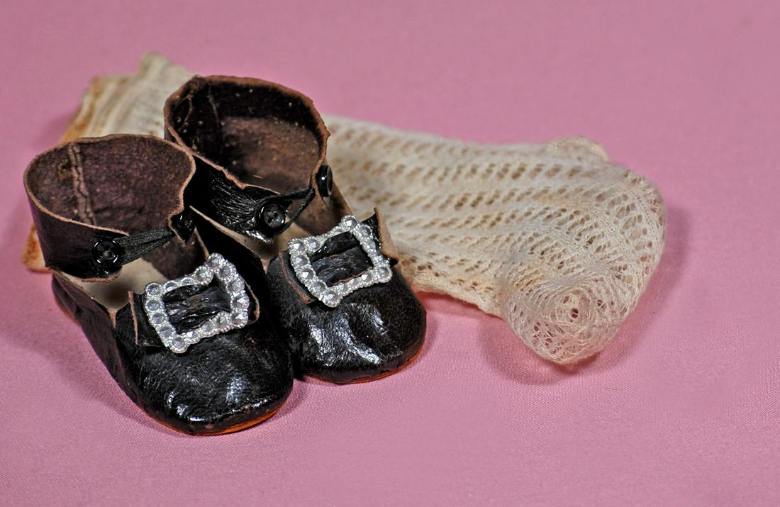 12. PAIR OF ANTIQUE DOLLS SHOES AND SOCKS. 2.5” L.: 12. PAIR OF ANTIQUE DOLLS SHOES AND SOCKS. 2.5” L. pair of soft black leather shoes with leather bows, silver buckle, ankle strap closing; plus pair of cream woven cotton socks. Commentary: Antique 