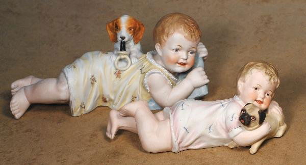 TWO GERMAN BISQUE PIANO BABIES WITH PUPPY. (1)10: TWO GERMAN BISQUE PIANO BABIES WITH PUPPY. (1)10 ½" x 5". All-bisque baby has sculpted brown hair, intaglio eyes and open/closed mouth with square-cut upper teeth, yellow floral dress, and blue pillo