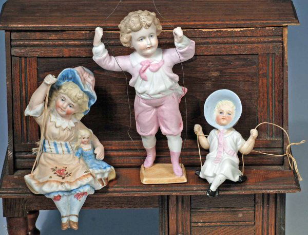 THREE GERMAN ALL-BISQUE SWINGERS. Includes: 5 ¾": THREE GERMAN ALL-BISQUE SWINGERS. Includes: 5 ¾" Girl holding doll, 6 ¾" boy in pink suit and boots, 4" girl in blue bonnet. Each is all-bisque with painted hair, features and costume; posed on bisq