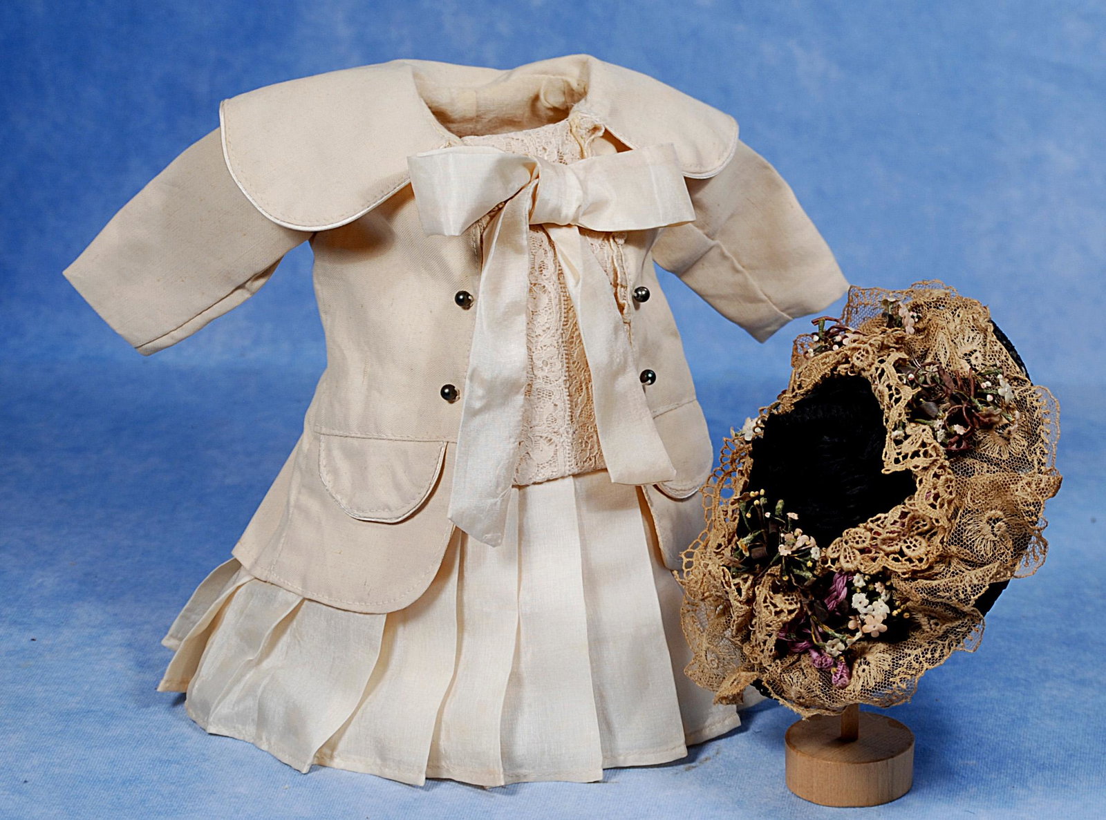 DOLL DRESS AND BONNET: DOLL DRESS AND BONNET. Cream silk jacket-style dress with lace front panel, pleated skirt, open front jacket with fitted and pleated back, 5” shoulder width, 10” length; plus black straw b