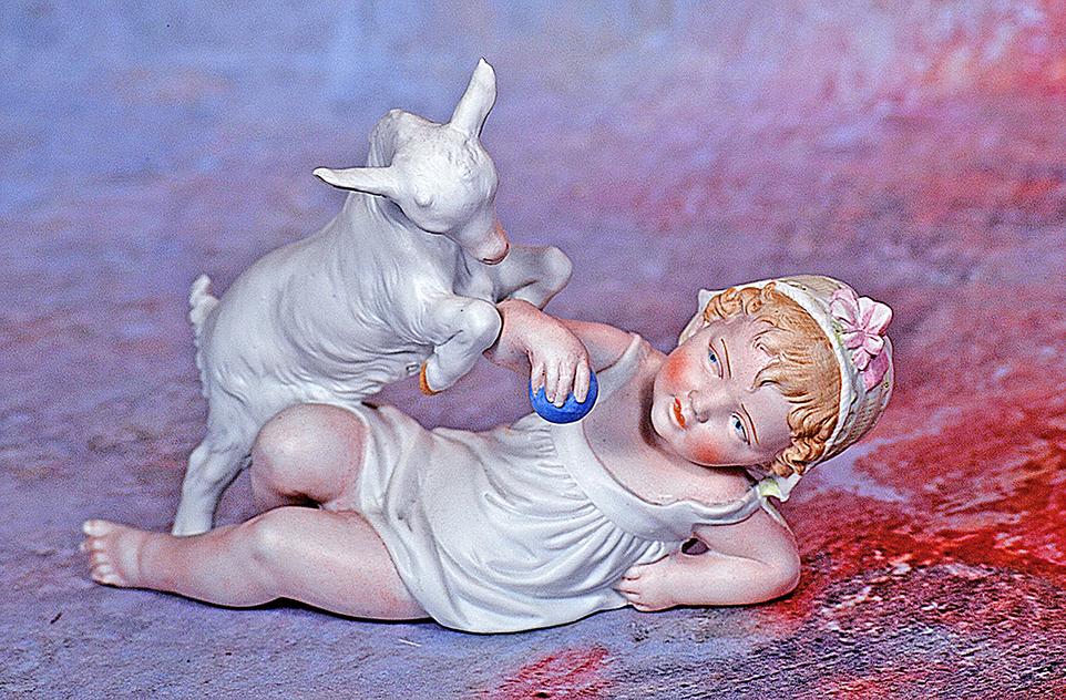 GERMAN BISQUE FIGURE OF GOAT PLAYING WITH BABY: 165. GERMAN BISQUE FIGURE OF GOAT PLAYING WITH BABY. 9” x 6 ½”. Marks: 9111. Bisque figure of baby with sculpted blonde hair and white bonnet having green ribbon and pink bow trim, pa