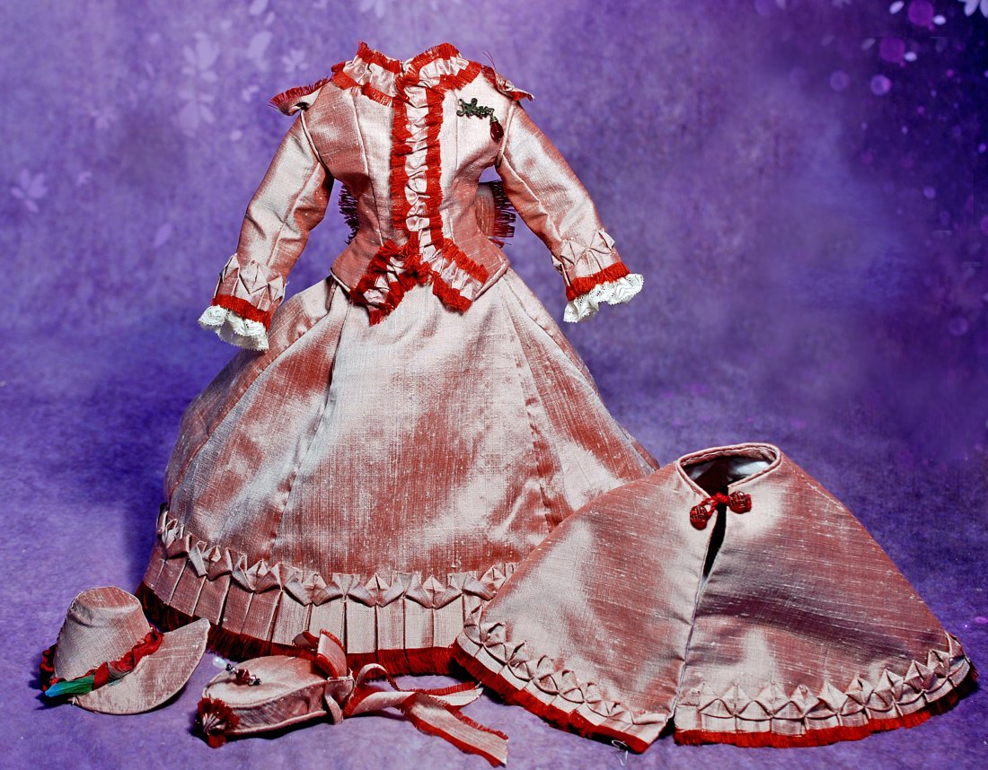 SILK OUTFIT FOR FASHION DOLL: SILK OUTFIT FOR FASHION DOLL. Rose silk two-piece gown with fitted bodice and skirt with pleated detail, matching cape, two bonnets, plus petticoat, all generously trimmed with maroon fringe trim. Siz