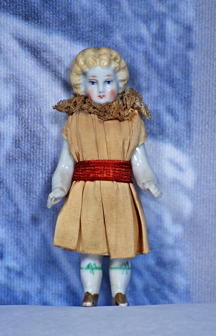 MINIATURE ALL-PORCELAIN DOLL WITH GILT BOOTS 4”: 4” Sculpted one-piece porcelain doll with sculpted blonde wavy hair, painted features, painted gilt boots and green ribbon accents below the knees, original dress Commentary
