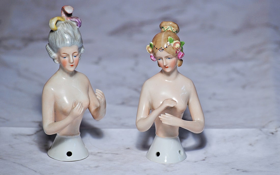 TWO GERMAN PORCELAIN HALF DOLLS BY GOEBEL: 4 ½” and 5” Marks: Goebel trademark symbol Each is waist-up, pink-toned porcelain nude figure of adult lady, posed with both arms away from body, painted features, downward glancing blue eyes, ta