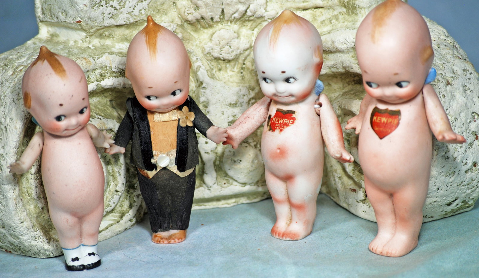 FOUR BISQUE KEWPIES: FOUR BISQUE KEWPIES. 4 ½” to 5 ½”. Each is all-bisque with classic Kewpie painted features, hair and wings, jointed at shoulders. One Kewpie in original black crepe paper formal