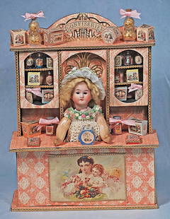 MINIATURE CONFISERIE WITH BISQUE DOLL AS CANDY BOX: MINIATURE CONFISERIE WITH BISQUE DOLL AS CANDY BOX. 12” x 9”. Heavy board storefront with shelved etagere arrangement containing many miniature candy boxes, assorted glass candy bottles an