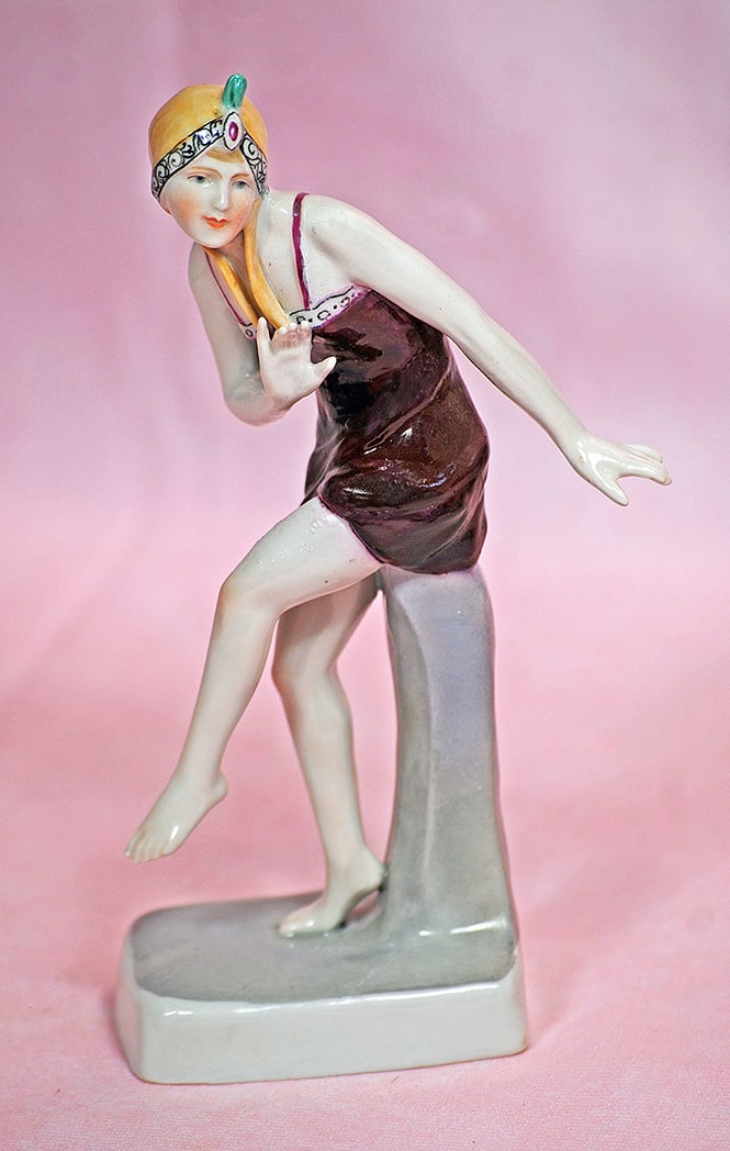 GERMAN PORCELAIN FLAPPER LADY IN STYLISH COSTUME: GERMAN PORCELAIN FLAPPER LADY IN STYLISH COSTUME. 6 ¾”. All-porcelain lady posed on self base has sculpted dress and bonnet with decorated details, painted features, blue eyes, tapered brow