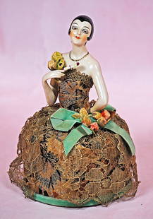 GERMAN PORCELAIN FLAPPER HALF DOLL AS PINCUSHION: GERMAN PORCELAIN FLAPPER HALF DOLL AS PINCUSHION. 5” doll; 9” overall. Waist-up pink-toned porcelain figure of flapper lady with sculpted black hair, sculpted necklace and earrings,