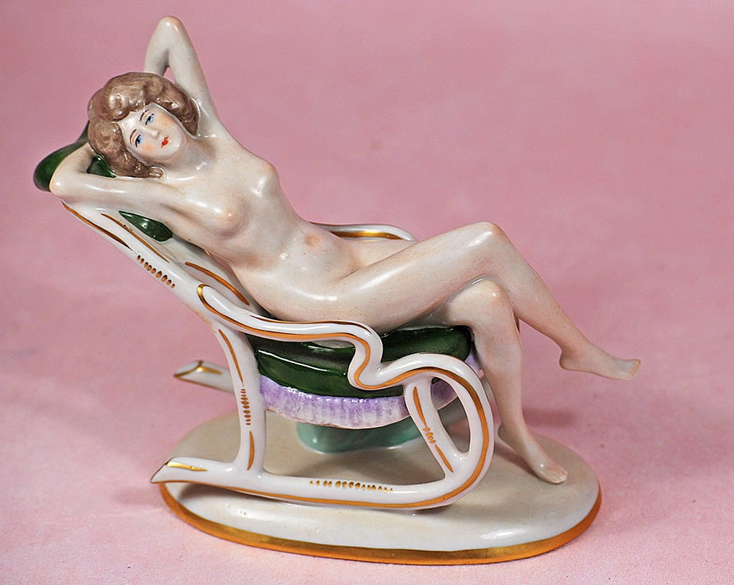 GERMAN PORCELAIN NUDE LADY IN ROCKING CHAIR BY SCHIEBE-ALSBAC: GERMAN PORCELAIN NUDE LADY IN ROCKING CHAIR BY SCHIEBE-ALSBAC. 6”. Marks: Blue trademark symbol. All-porcelain figurine of nude lady with sculpted brown hair and blue eyes is posed with arms beh