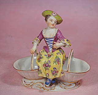 GERMAN PORCELAIN FIGURINE OF YOUNG CHILD POSED ON BASKET: GERMAN PORCELAIN FIGURINE OF YOUNG CHILD POSED ON BASKET. 6&#8221;. Marks: Blue V stamp. All-porcelain figure of young child posed atop oval-shaped basket with twin handles and gilt decorations; sculp