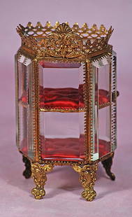 GILDED BRONZE MINIATURE DISPLAY CABINET: GILDED BRONZE MINIATURE DISPLAY CABINET. 6”. Gilded ormolu display cabinet with beveled edge windows on the door, top and two sides, has tufted red silk shelves, ormolu feet and fancy crest. 